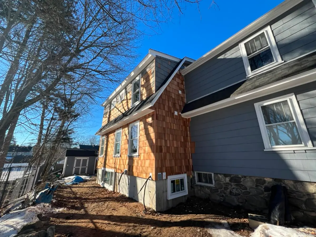 Cedar-shingled addition in Reading, MA above its fieldstone foundation, beside the grey main house